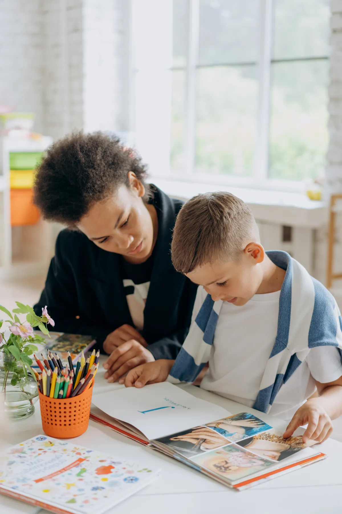 An adult helps a boy read a book at a learning table.