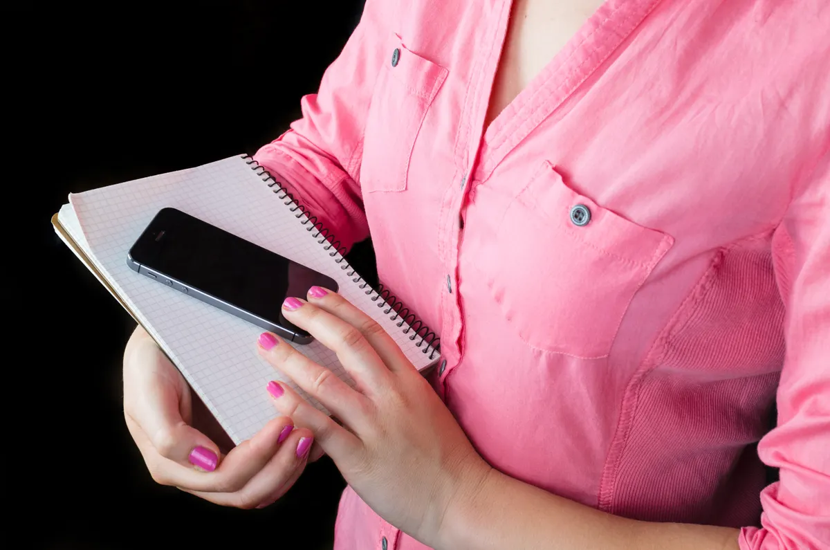 A person holds a phone and notebook while keeping notes.