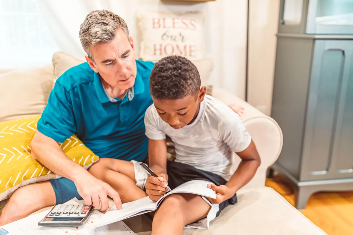 A child writes in a workbook while an adult helps him follow the lesson.