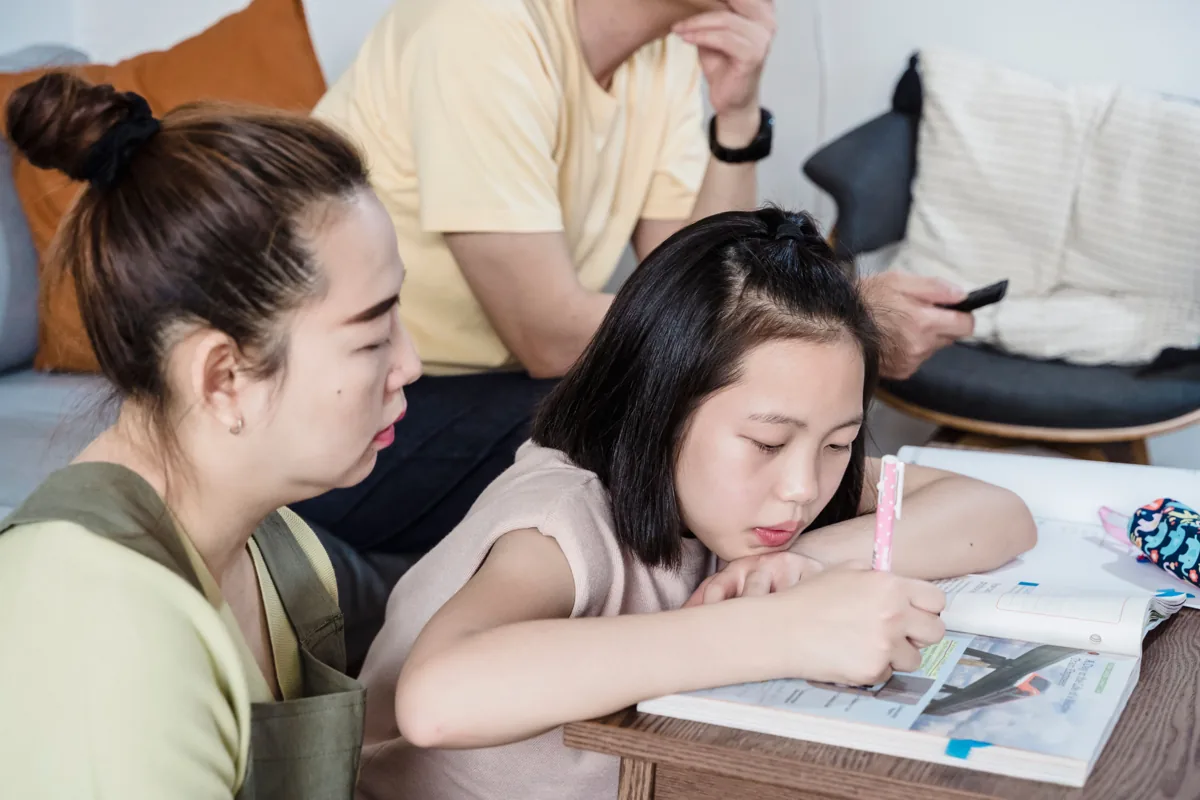 A mother sits beside her daughter as she reads and works in a workbook.