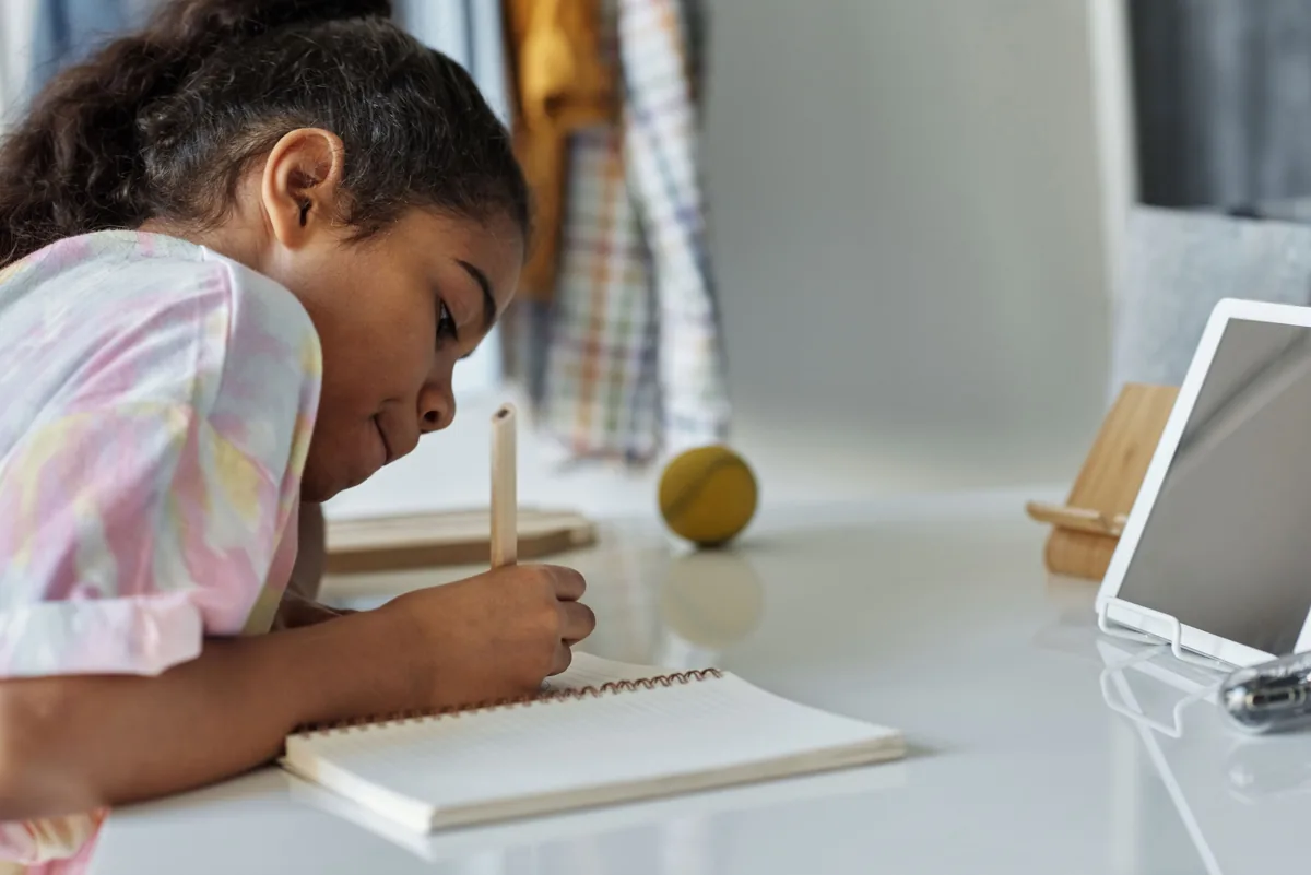 A girl practices writing in a notebook beside a tablet.