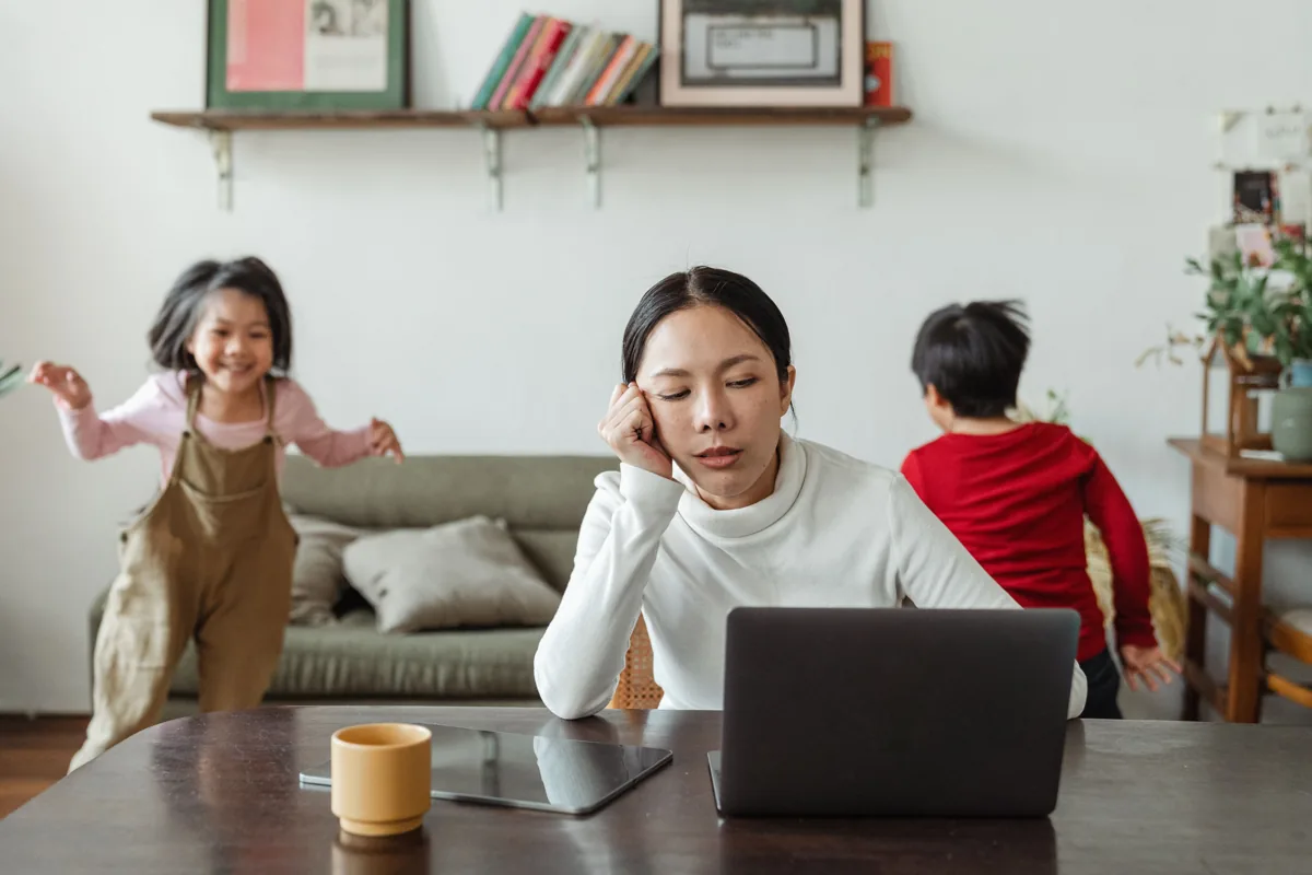 A mother rests her head on one hand at her laptop while her children play nearby.