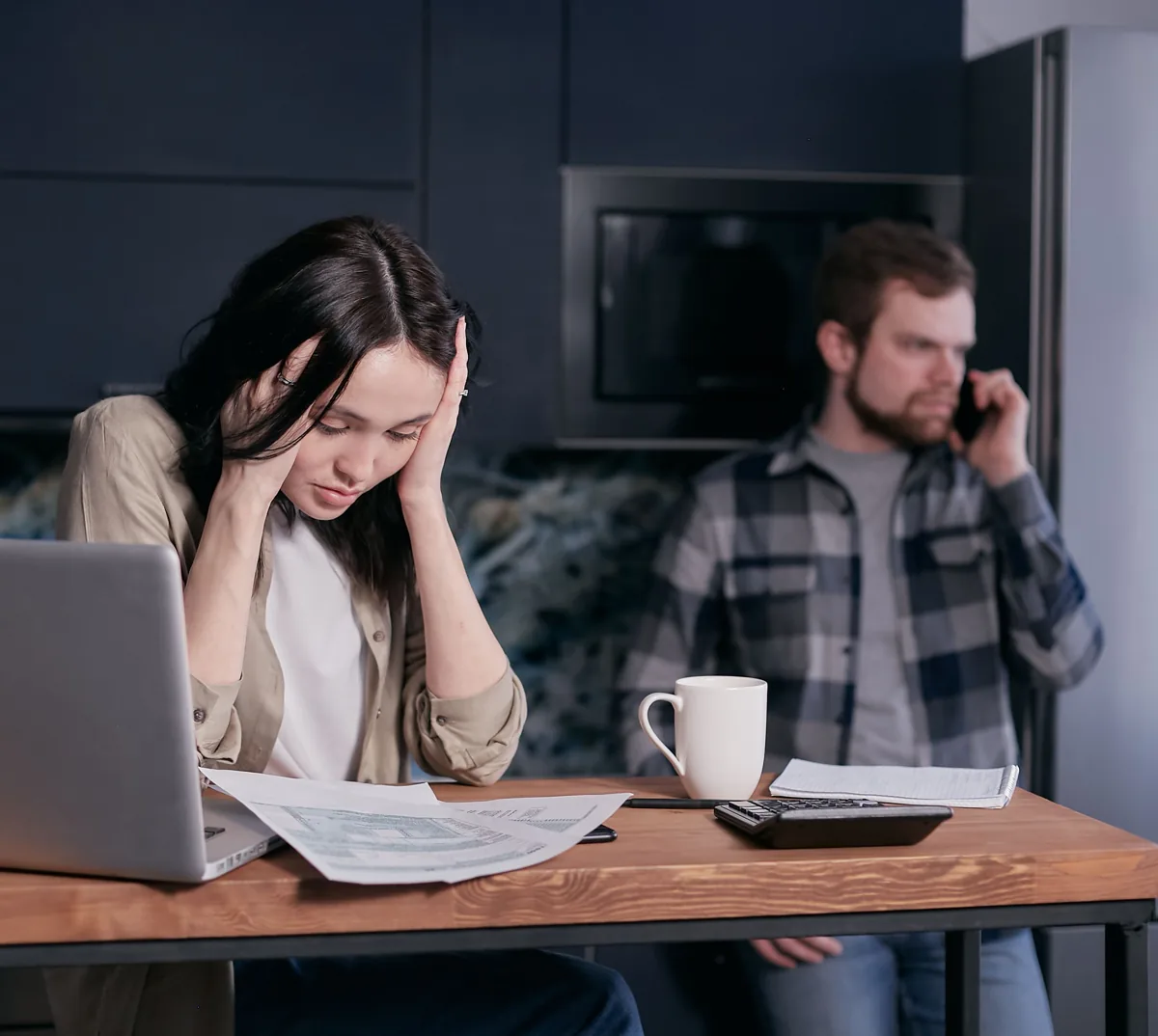 An adult holds her head over paperwork while another adult talks on the phone beside her.